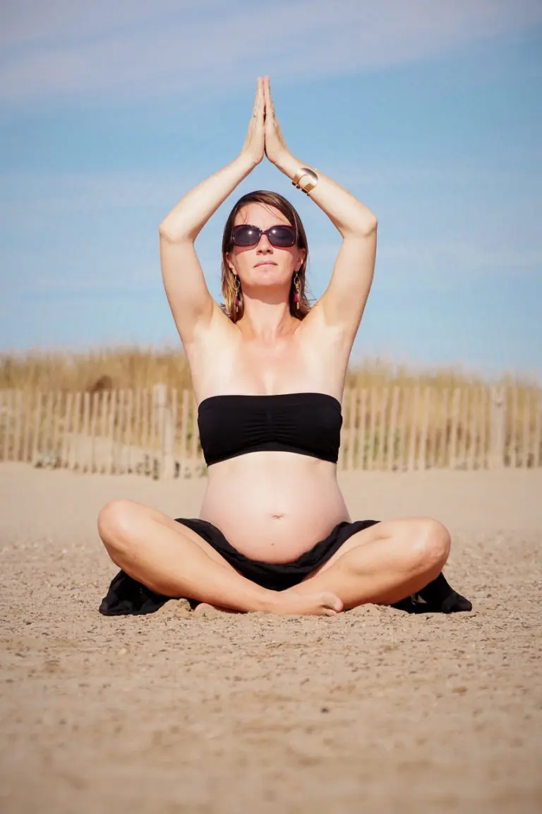 future maman en posture de yoga ventre nu sur la plage lunettes de soleil palissade bambou