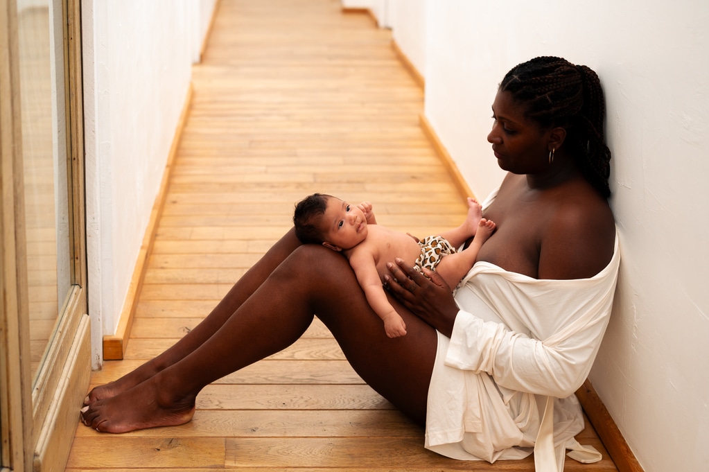 photo d'une femme assiste dans un couloir sur du beau parquet, vétue d'un peignoir, son nouveau né sur ses genoux. Photo prise par LONOWAÏ Photographie lors d'une séance photo de maternité intimiste à Narbonne