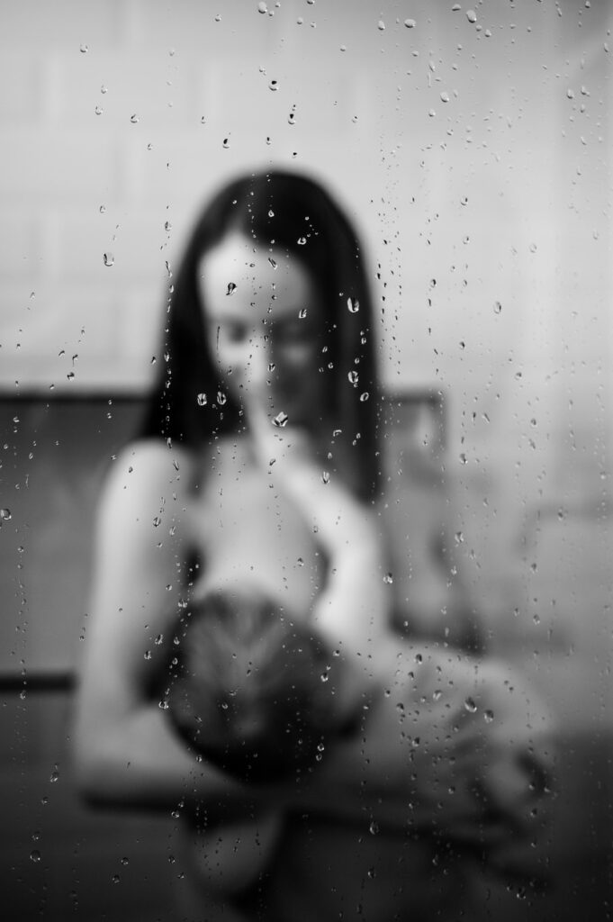 femme avec un bebe dans les bras en peau à peau sous la douche vue à travers une vitre couverte de gouttes d'eau. Photo prise lors d'une séance de maternité intimiste à Narbonne par LONOWAÏ Photographie